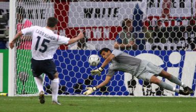 Two soccer players on a field, one attempting to score by kicking the ball towards the goal while the goalkeeper is diving to block the shot. The field is bordered by a blue and white wall with a logo and spectators in the background.