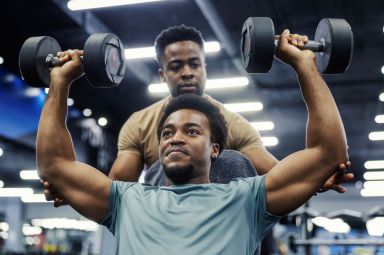 Two young black men training in a gym, one lifting dumbbells while his personal trainer spots and coaches him during workout