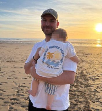 An individual, wearing a cap and a white t-shirt, is holding a baby in their arms on a beach, with a setting sun in the background.