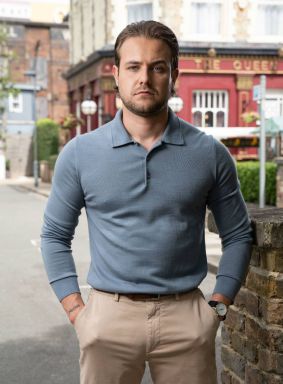 An individual dressed in a dark blue polo shirt stands on a city street, with a vibrant red building and various urban elements in the background.