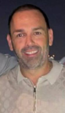 A close-up portrait of an individual with a clean-shaven face, sporting a warm, broad smile and exuding an air of contentment. The person is dressed in a gray shirt, and their expression is framed by short, neatly trimmed hair.