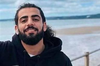 An individual with a beard, dressed in a black hoodie, is standing on a coastal pathway, with the sea visible in the background. The sky above is partly cloudy.
