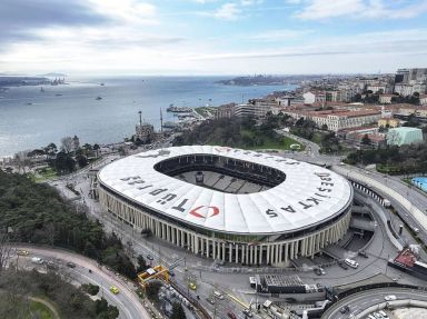 An aerial view of a modern, large-capacity stadium, nestled alongside a coastal city with urban infrastructure and a serene seascape in the background.