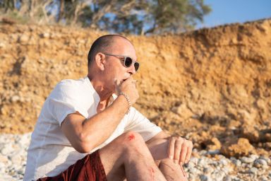 A man in his fifties smoking on a pebble beach at sunset on a summer day. 