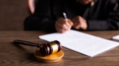 A wooden gavel rests on top of a wooden desk. In the background, out of focus is someone writing on a note pad- we can't see their face.
